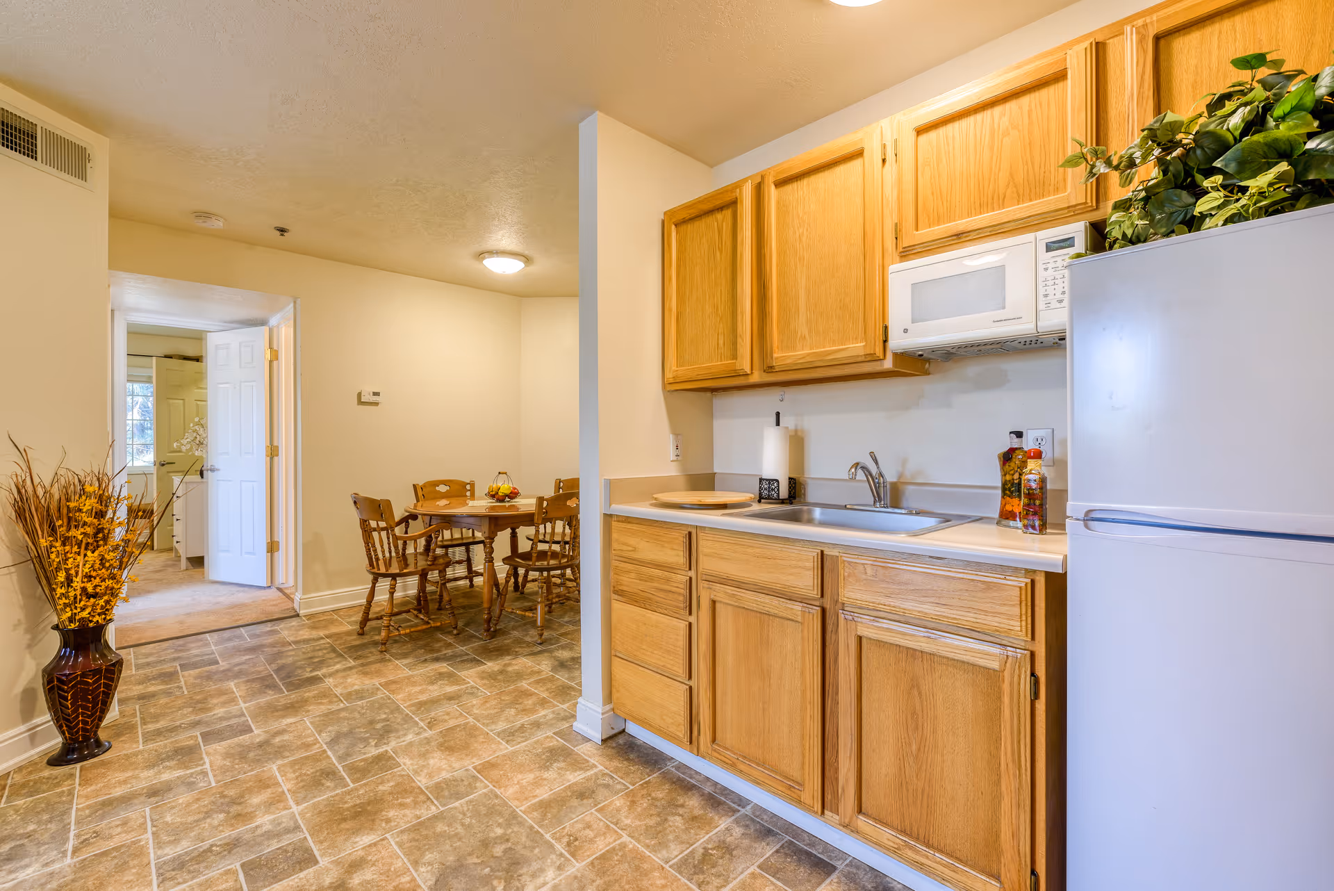 Small apartment kitchen with oak cabinets, a sink and microwave beside a white refrigerator, and a wooden dining table visible in the background.