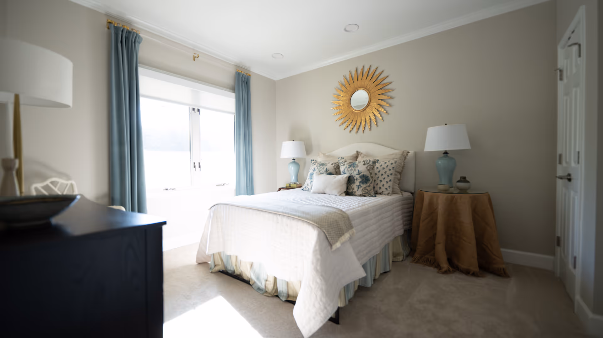 Bright, well-decorated bedroom with a bed, bedside tables and lamps, a large window with blue curtains, and a sunburst mirror above the headboard.