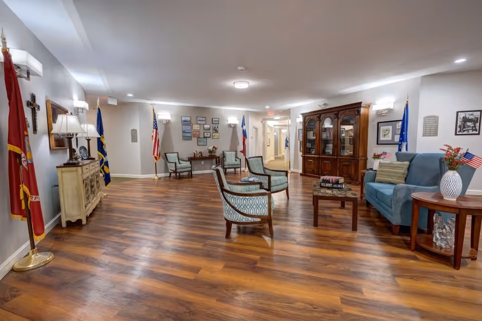 A spacious and well-lit common area with wooden flooring, several upholstered chairs arranged around a coffee table, a blue sofa, and a wooden cabinet with glass doors. The room is decorated with flags, framed pictures, a cross on the wall, and a side table with a vase of flowers and small American flags.
