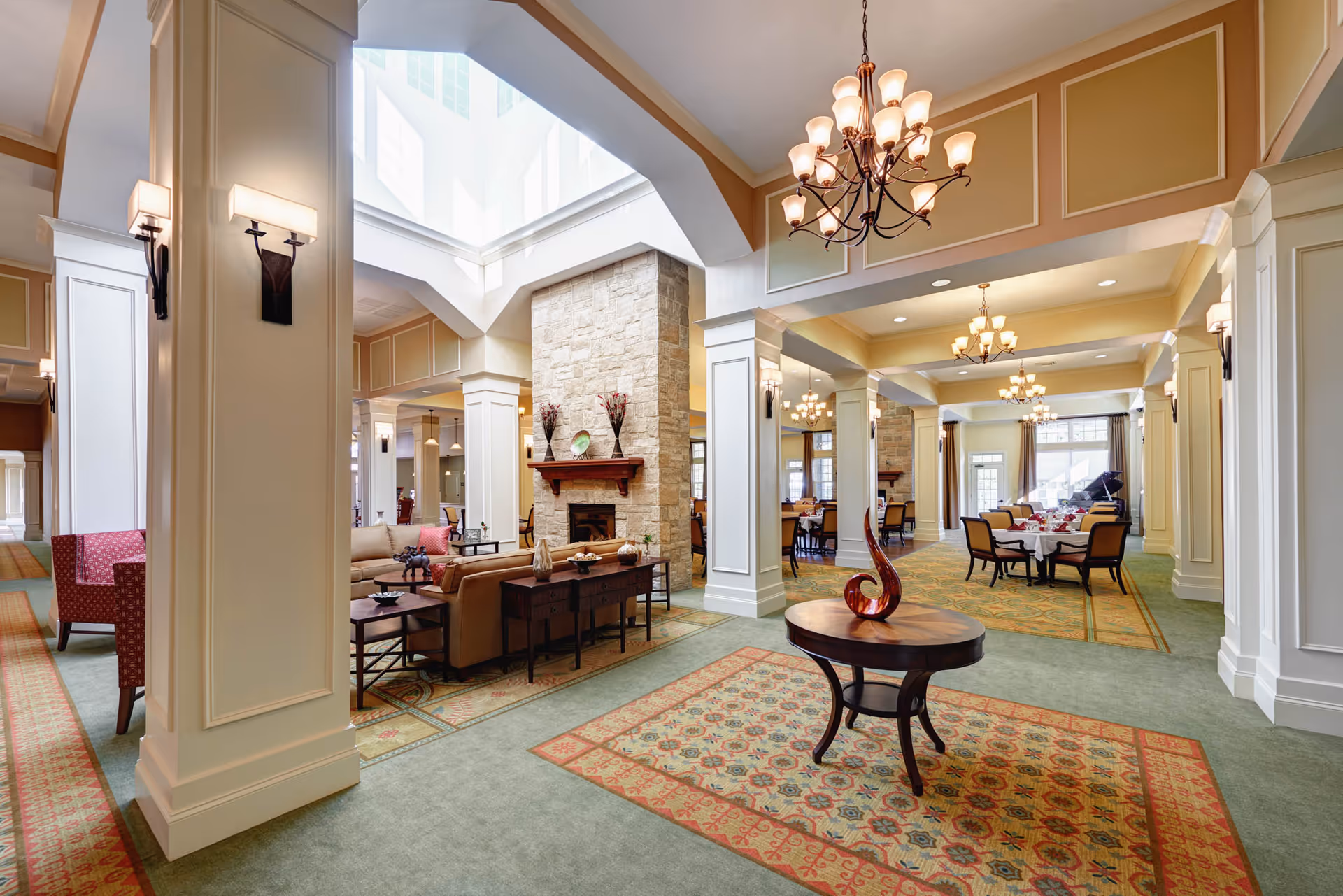 Spacious and elegant common area in a senior living facility featuring high ceilings with chandeliers, a stone fireplace, comfortable seating arrangements including sofas and armchairs, decorative rugs, and a round wooden table with a modern sculpture in the center.