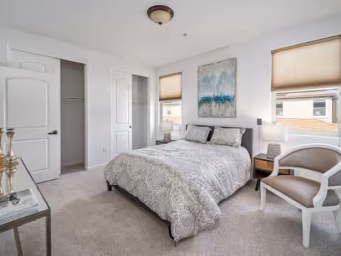 A bright bedroom with a neatly made bed featuring patterned bedding. The room has two windows with beige blinds, a painting above the bed, two bedside tables with lamps, a white armchair with a cushion, and an open closet door. The walls and carpet are light-colored, creating a clean and cozy atmosphere.