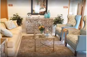 A cozy living room area with a beige sofa and two patterned armchairs around a glass coffee table. The room features a decorative cabinet with plants and vases on top, a large mirror on the wall, and a soft area rug covering the floor.