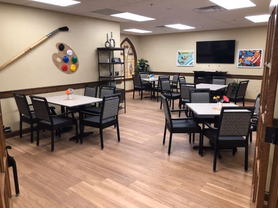 A well-lit room with several square tables, each surrounded by four chairs. The room has light-colored walls and wood flooring. On the walls are decorative items including a large artist's palette with colorful paint blobs, a wooden oar, and framed pictures. A flat-screen TV is mounted on the far wall above a black cabinet. There is a black shelving unit with decorative items and a potted plant in the corner.