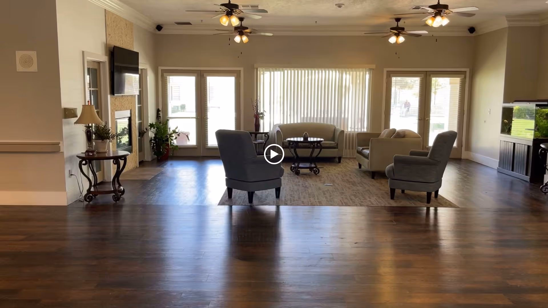 Bright communal living room with armchairs and sofas arranged around coffee tables on hardwood floors under ceiling fans and large windows.