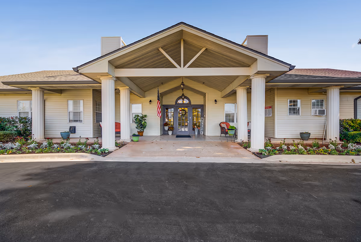 Front entrance of Hoyt Court facility with a covered porch supported by white columns, potted plants, an American flag, and a welcome sign near double glass doors.