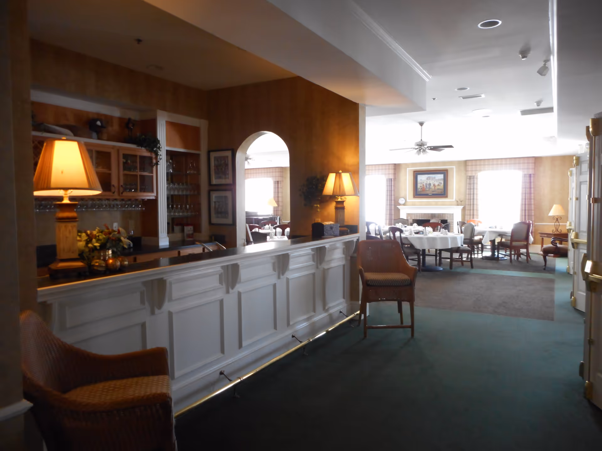 Interior view of a senior living facility dining area with a white paneled counter and two wicker chairs in the foreground. The counter has a lamp and decorative items on it. In the background, there are round dining tables with white tablecloths and chairs, a fireplace with a framed painting above it, and large windows with plaid curtains letting in natural light.