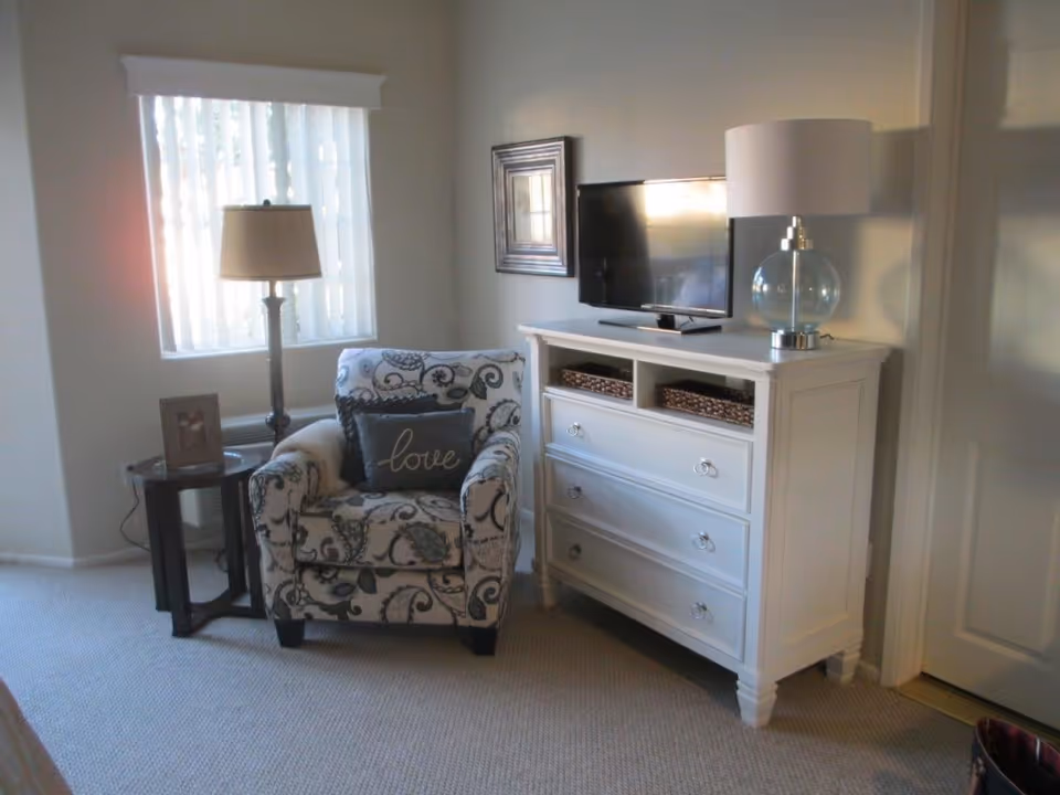 A cozy corner of a living room featuring a patterned armchair with a 'love' pillow, a small round side table with a framed photo, a floor lamp next to a window with vertical blinds, and a white dresser with a flat-screen TV and a glass table lamp on top.
