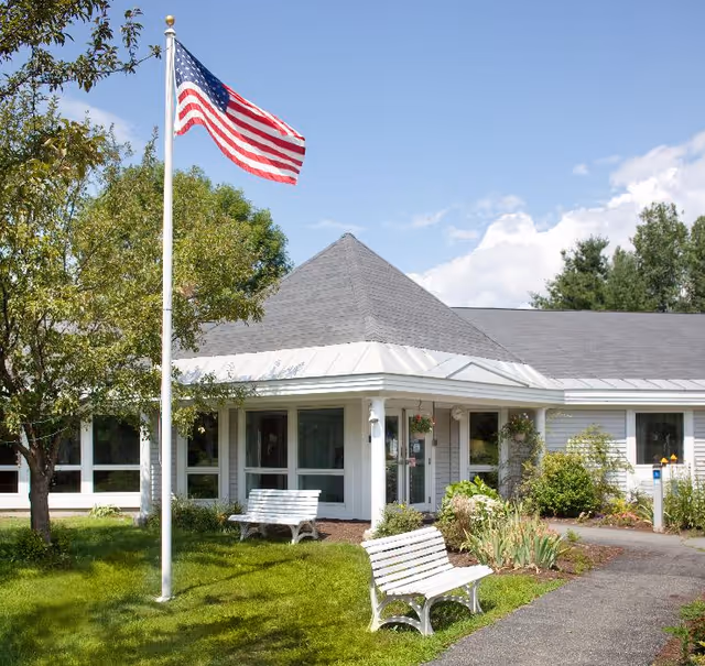 Exterior view of a single-story building with a gray roof and white siding, surrounded by green grass, trees, and plants. Two white benches are placed on the lawn near a paved walkway. An American flag on a flagpole is prominently displayed in front of the building under a partly cloudy blue sky.