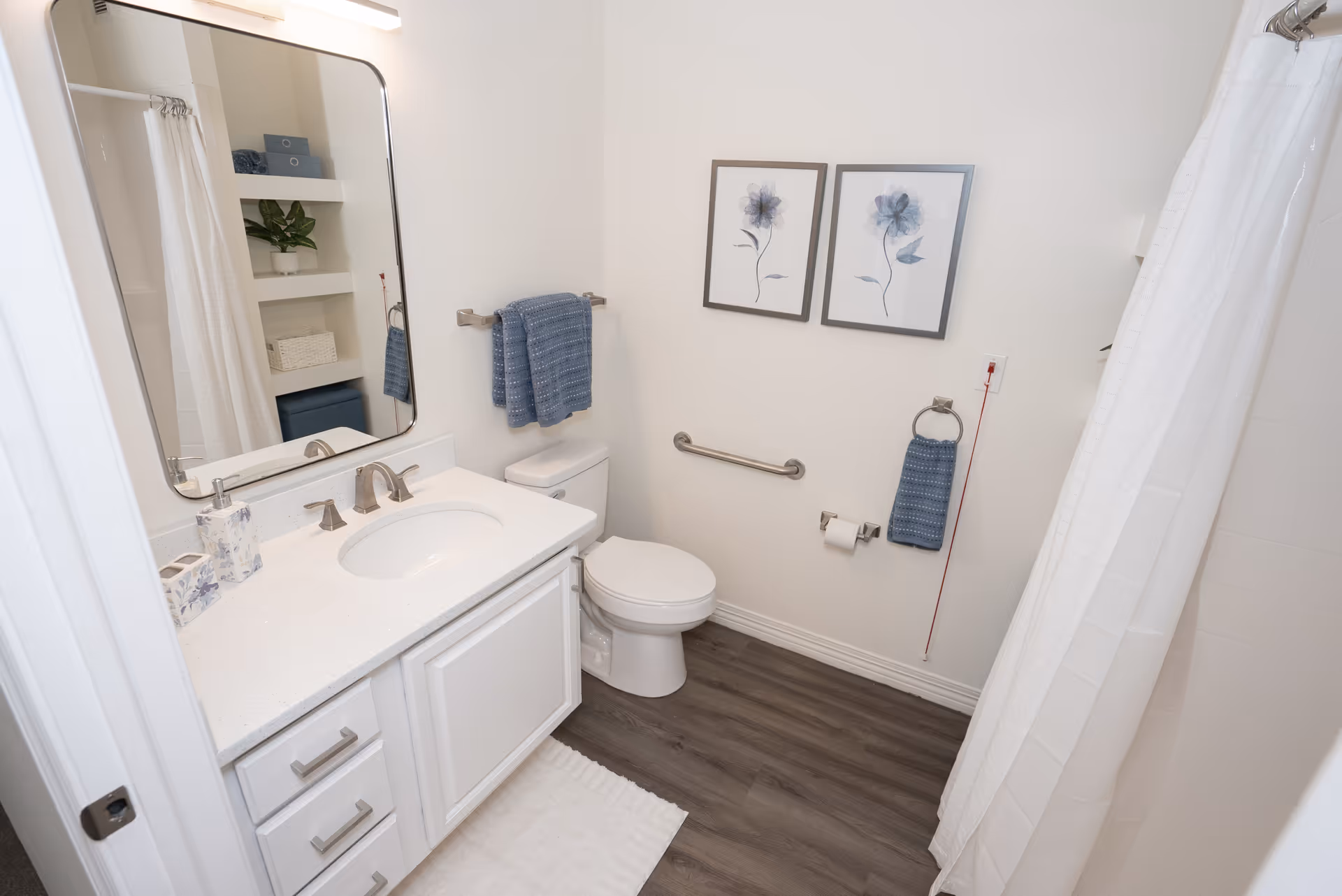 A clean and modern bathroom with a white vanity and sink, a large mirror above the sink, a toilet with a blue towel hanging on a rack above it, two framed floral artworks on the wall, a grab bar next to the toilet, a toilet paper holder with a roll, a hand towel ring with a blue towel, and a shower with a white curtain. The floor is wood-style laminate and there is a white bath mat in front of the vanity.