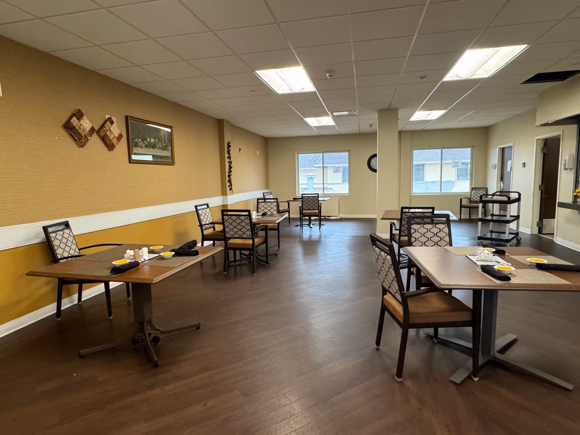 A dining room in a senior living facility with several tables and chairs arranged neatly. Each table has place settings including plates, napkins, and utensils. The room has a warm color scheme with brown flooring, beige walls, and decorative wall art. Large windows let in natural light, and there is a clock on the far wall.