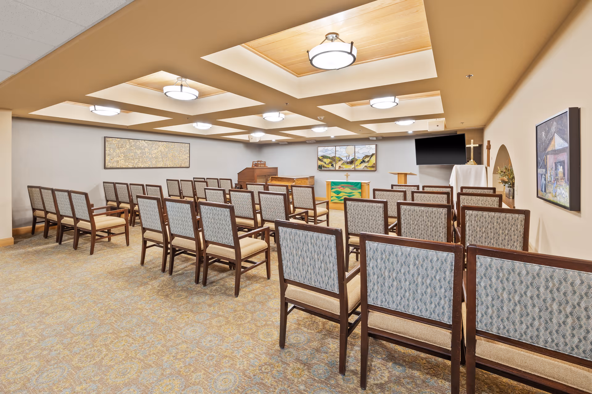 A small chapel-style meeting room with rows of upholstered chairs facing an altar area, piano, and wall-mounted TV.