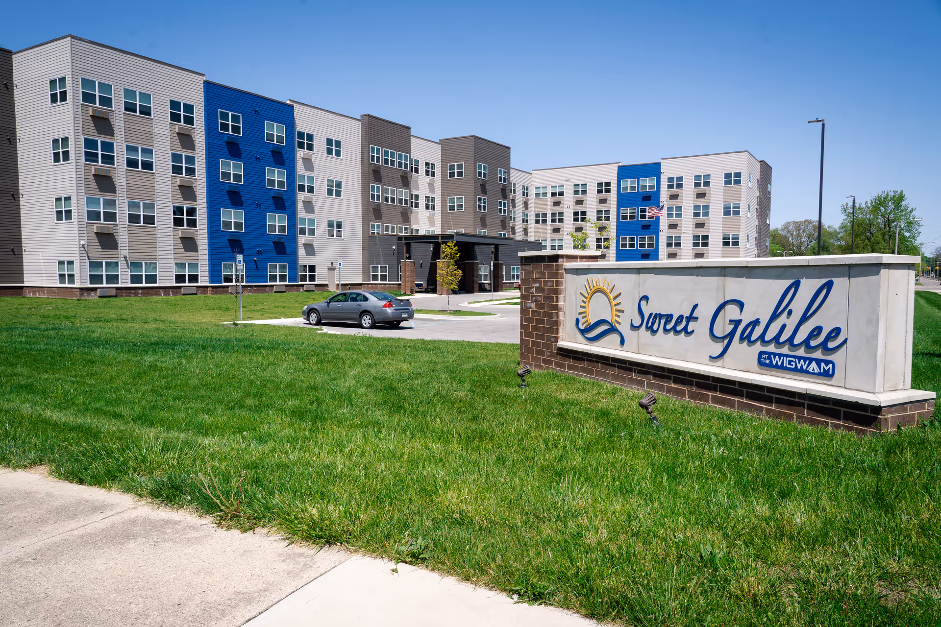Exterior view of Sweet Galilee at the Wigwam, a multi-story senior living facility with beige, blue, and gray siding. A large sign with the facility name is in the foreground on a grassy lawn, and a silver car is parked near the building entrance under a clear blue sky.