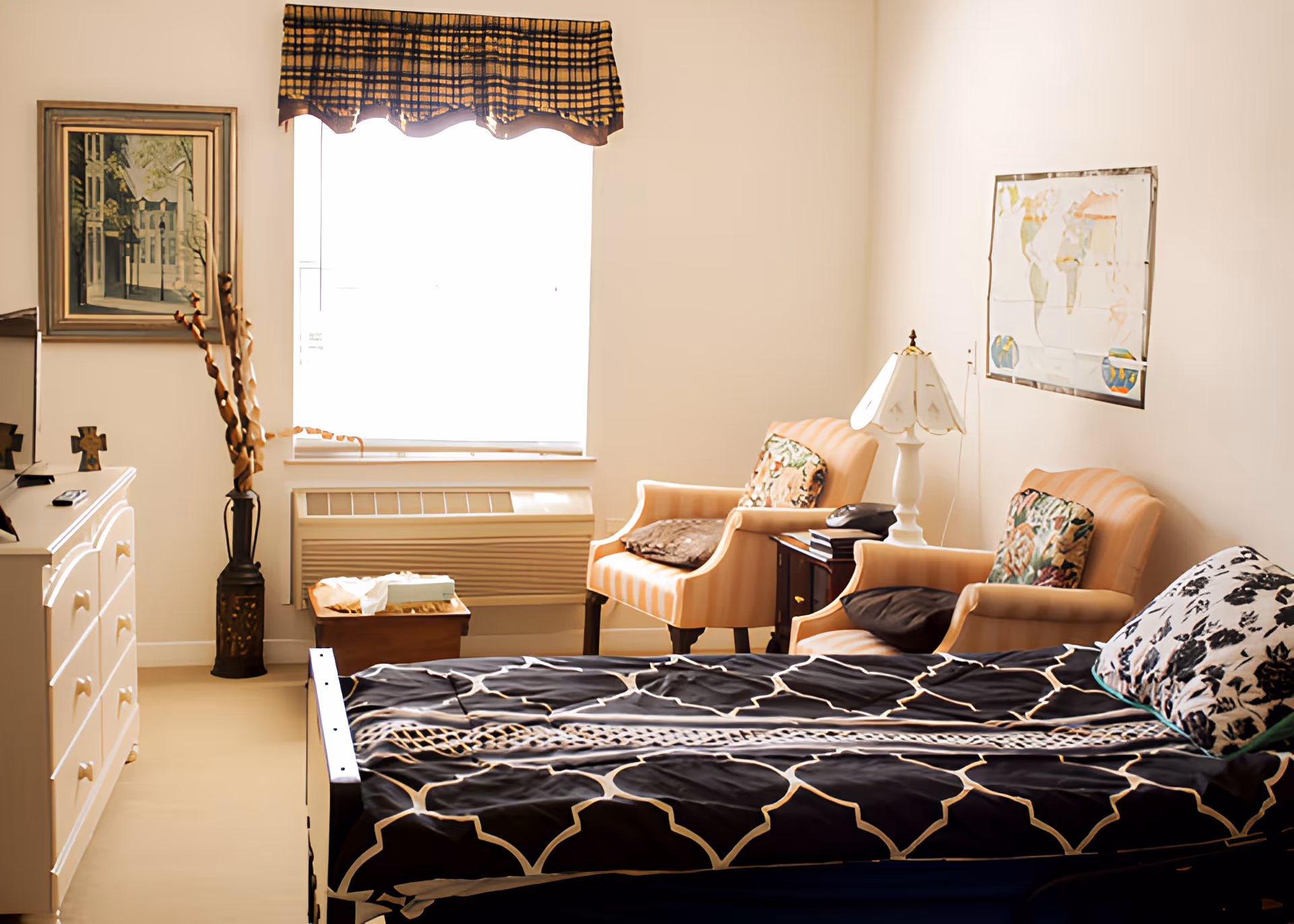 Sunlit assisted living bedroom with a single bed, two upholstered armchairs, a dresser, and a window with a valance.