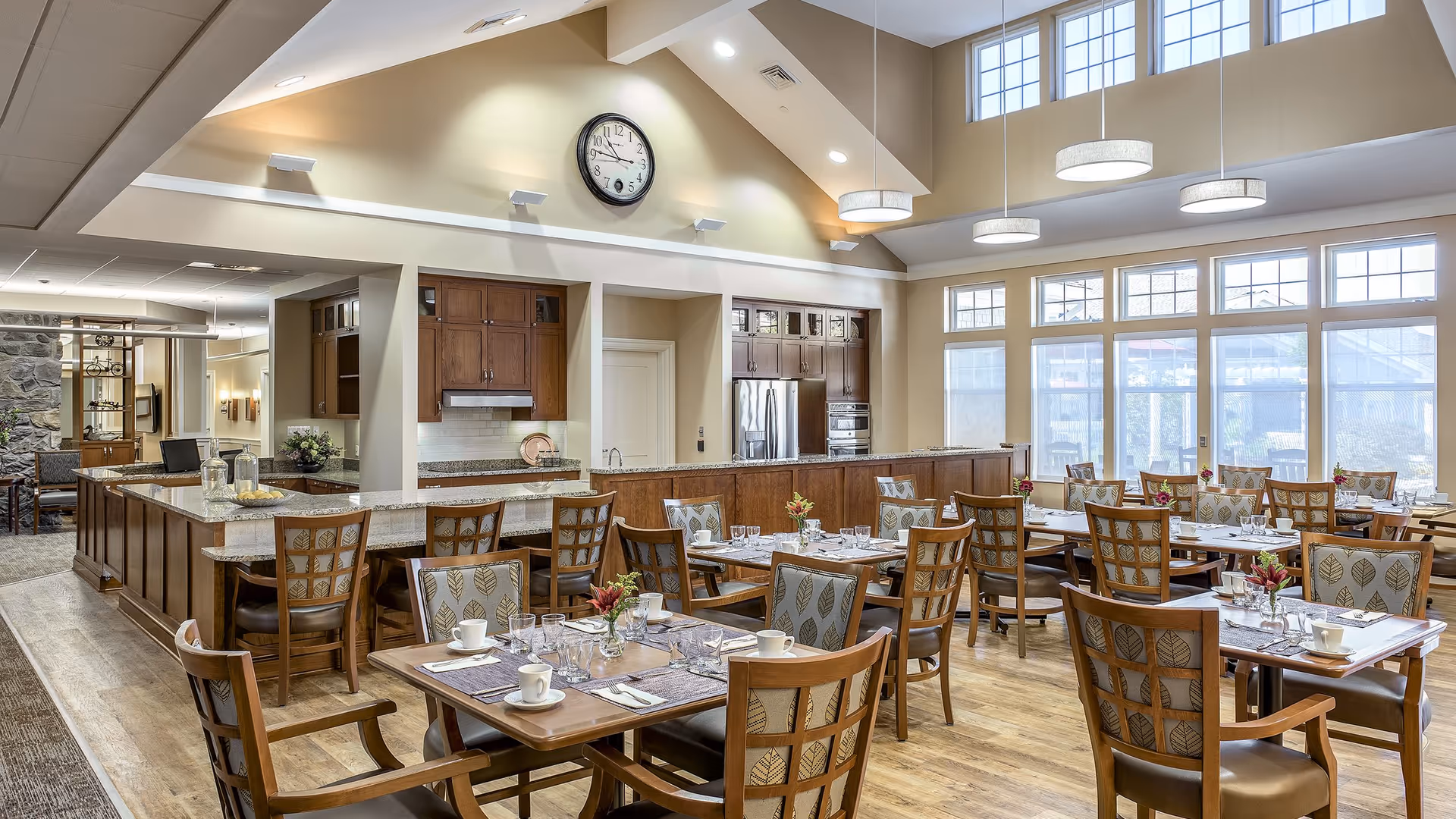 A bright and spacious dining room with multiple wooden tables and chairs arranged neatly. Each table is set with placemats, cups, glasses, and small flower arrangements. The room features large windows allowing natural light to fill the space, a high vaulted ceiling with modern pendant lights, and a kitchen area with wooden cabinetry and stainless steel appliances in the background. A clock is mounted on the wall above the kitchen area.