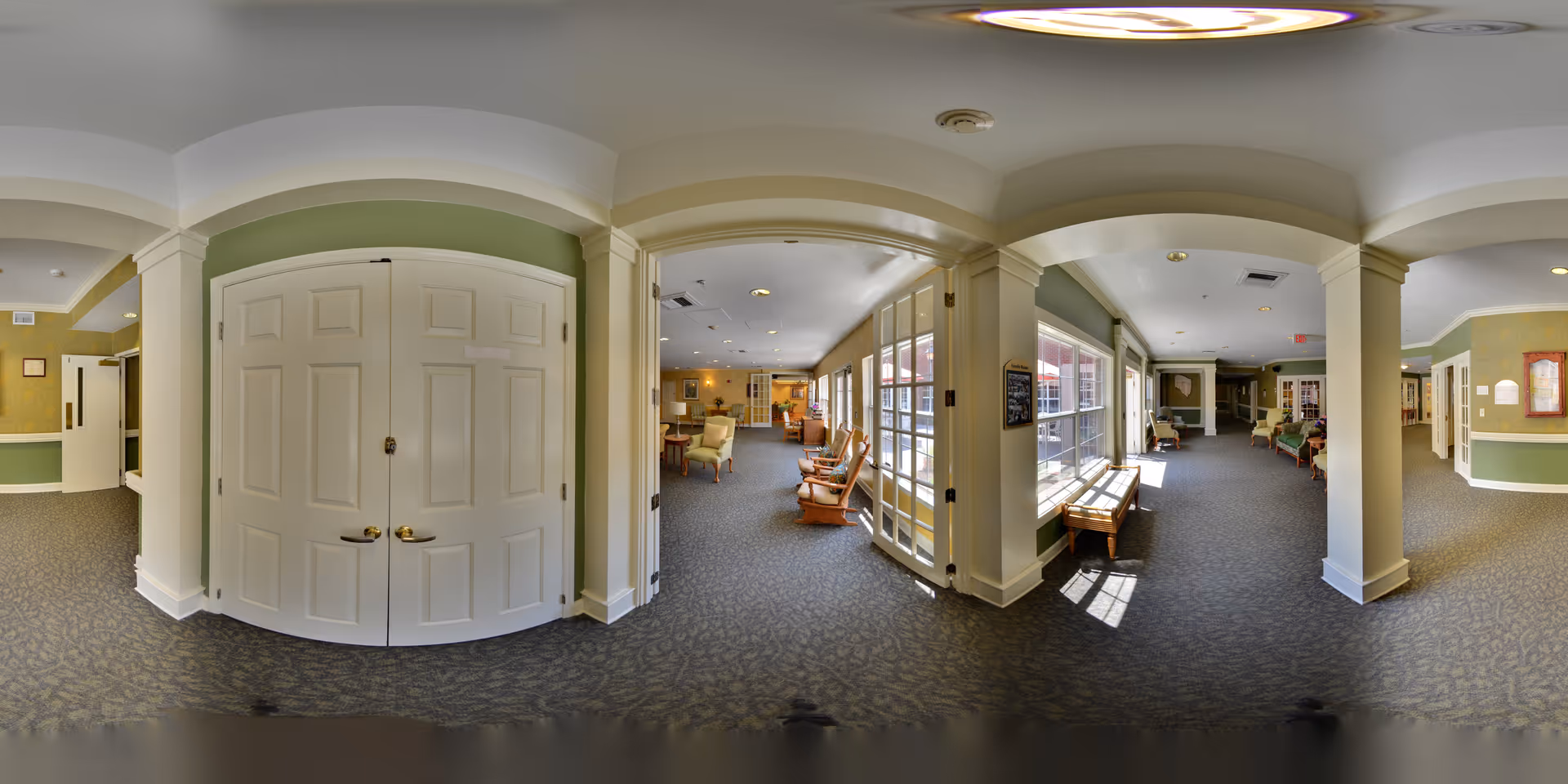 A spacious, well-lit common area in a senior living facility with carpeted floors, white pillars, and green walls. The area features multiple seating options including wooden rocking chairs and cushioned armchairs. Large windows allow natural light to fill the space, and there are double doors and bulletin boards visible on the walls.