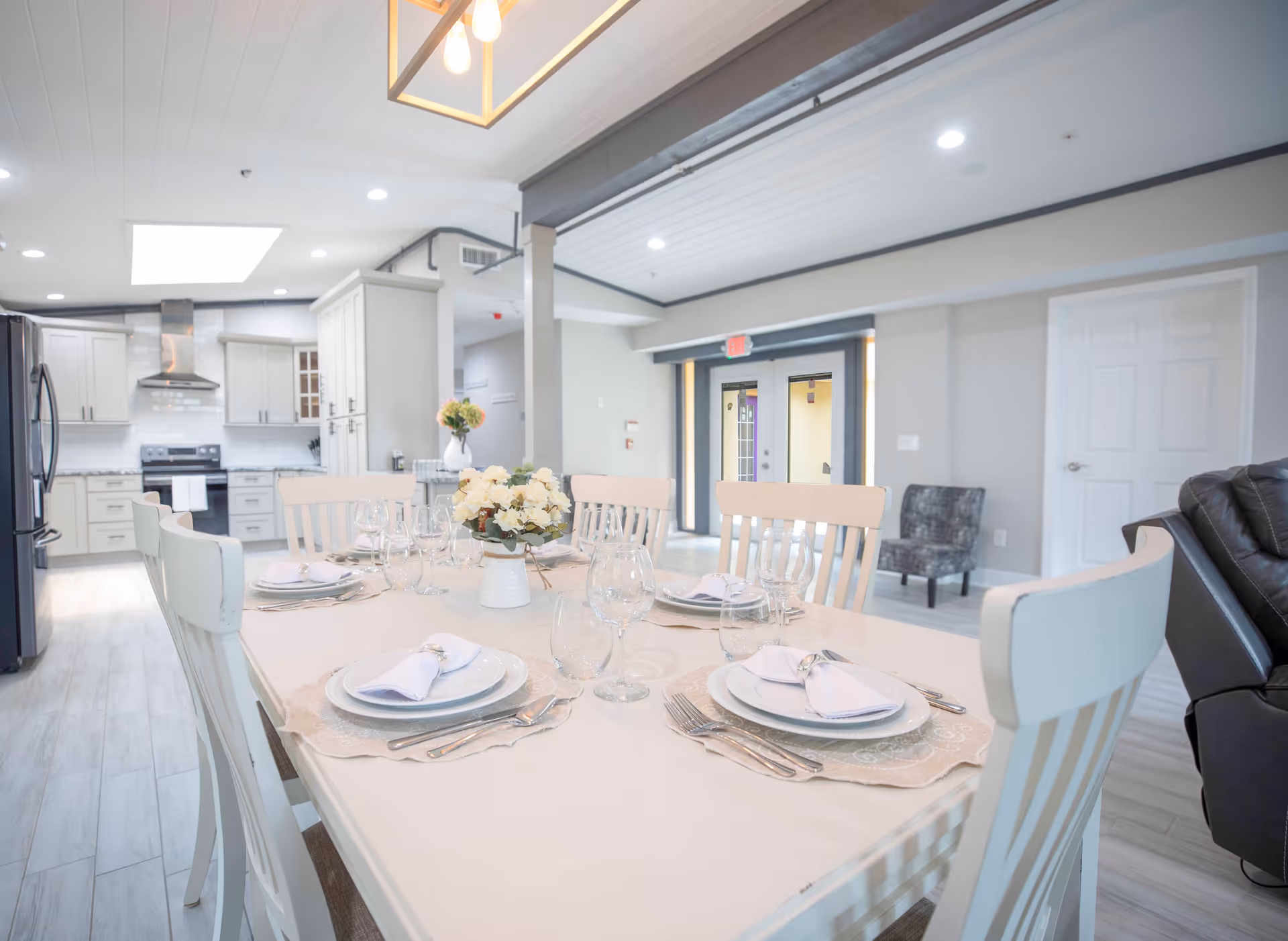 A bright and modern dining area with a white dining table set for six people, featuring plates, napkins, cutlery, and glassware. The table has a centerpiece with white flowers. In the background, there is a spacious kitchen with white cabinets, stainless steel appliances, and a range hood. The room has light-colored flooring and walls, recessed lighting, and a comfortable seating area with a chair and a sofa visible.