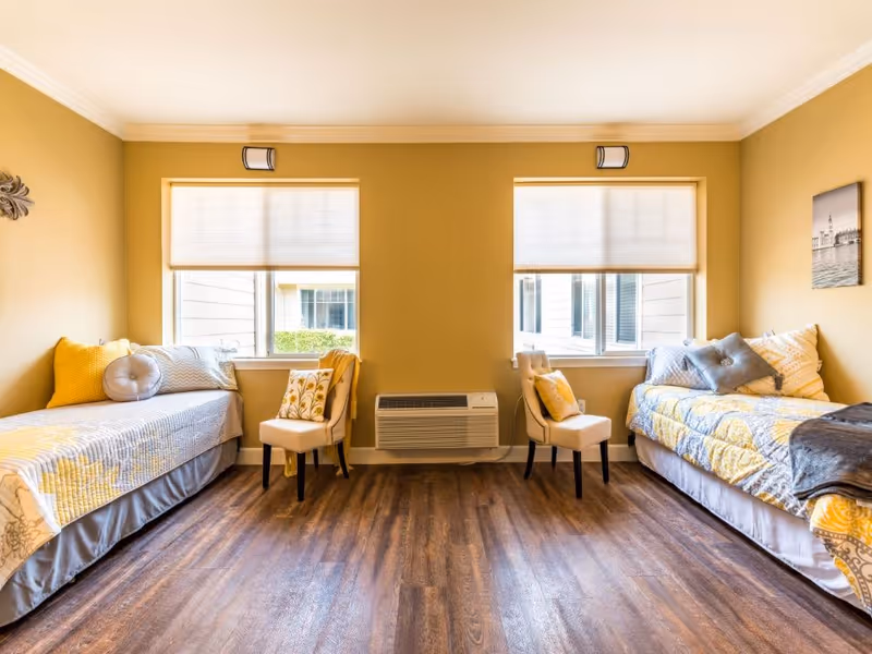 Bright shared bedroom with two twin beds, yellow-accented bedding, chairs, and windows.
