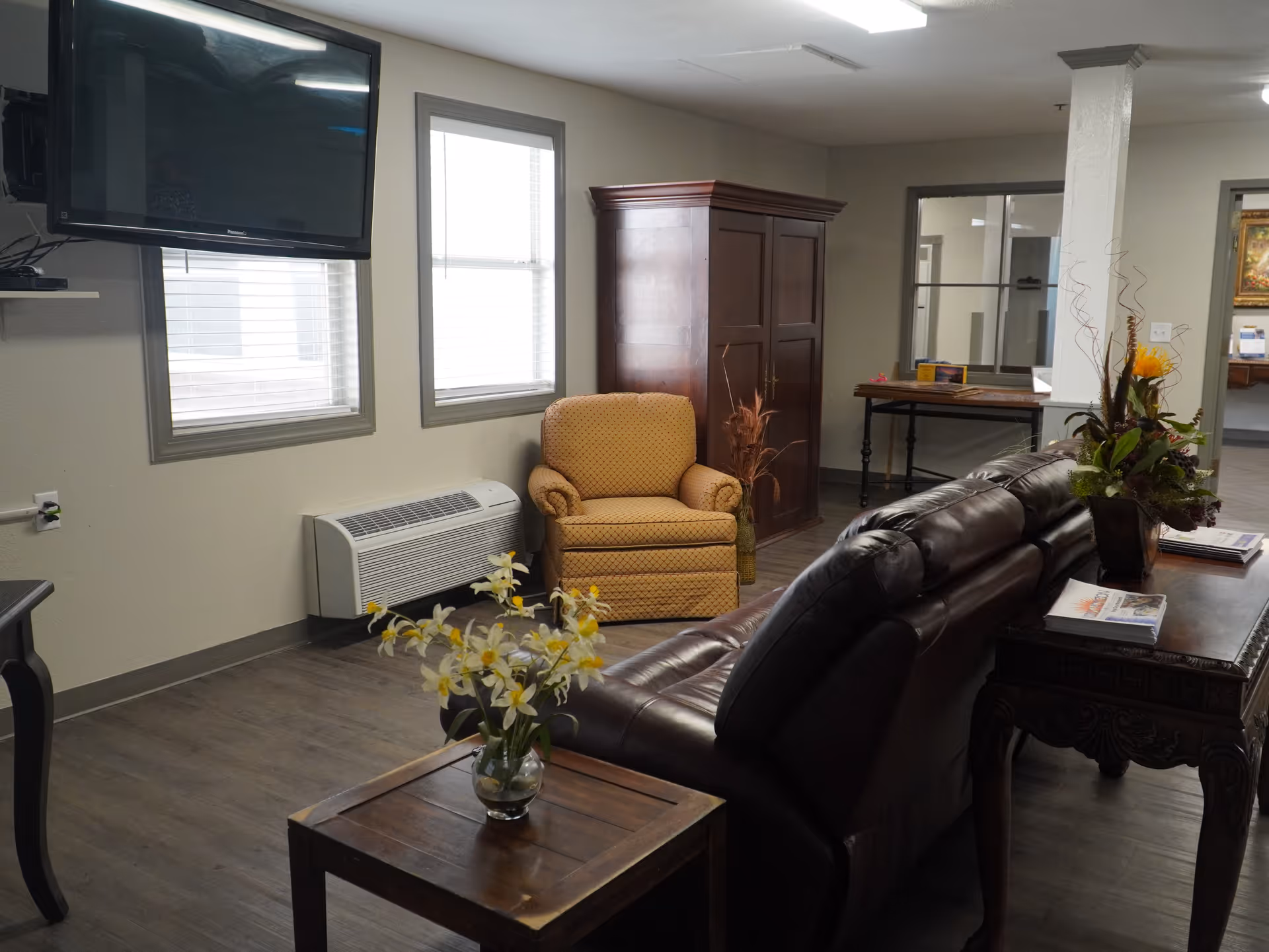 A cozy living room area with a brown leather sofa, a wooden side table with a vase of flowers, a yellow upholstered armchair, a wooden cabinet, and a wall-mounted flat-screen TV. The room has two windows with blinds and a wooden floor.