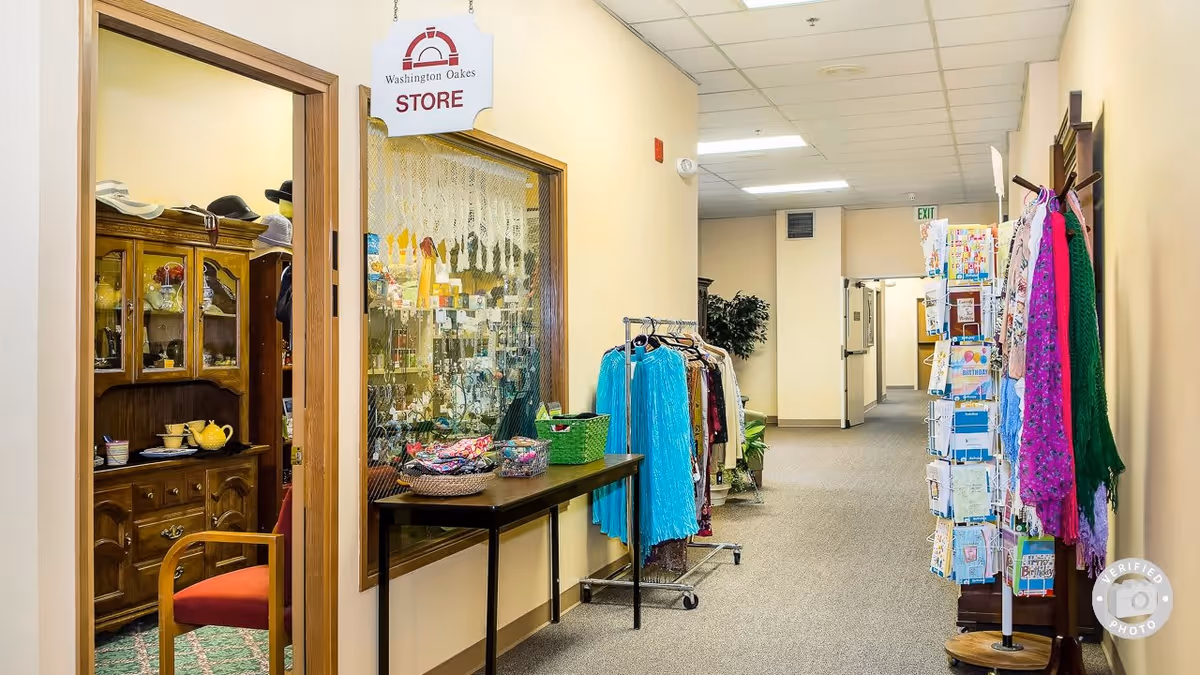 Interior hallway of Washington Oakes facility with a small store area visible through a window and open door. The store has shelves with various items including hats and teapots. In the hallway, there are racks with clothing and a rotating display stand with greeting cards. The hallway is well-lit with fluorescent ceiling lights and has beige walls and carpeted floor.