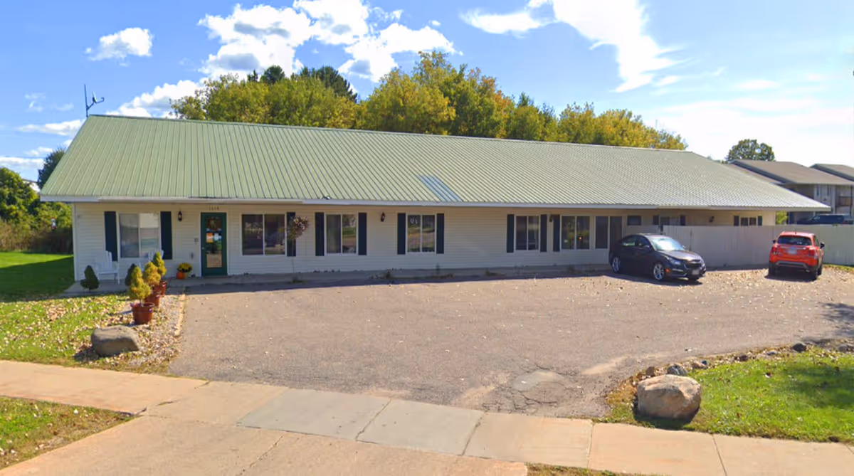 Single-story building with a green metal roof and white siding, surrounded by trees and a parking area with two cars parked. The building has several windows and a green door with potted plants near the entrance.