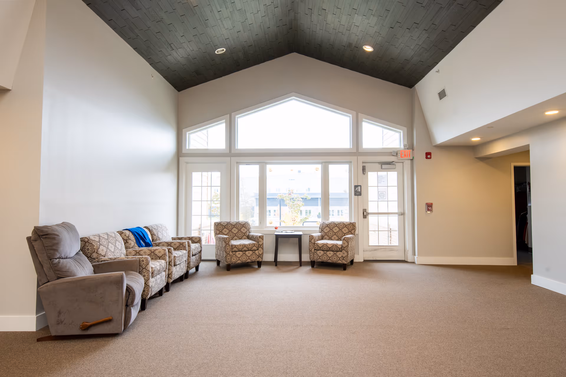 A bright and spacious common area with a high vaulted ceiling featuring dark wood paneling. The room has large windows and a glass door letting in natural light. There are five upholstered armchairs arranged along the walls, with a small table between two of the chairs near the windows. The walls are painted light beige, and the floor is carpeted in a neutral tone.