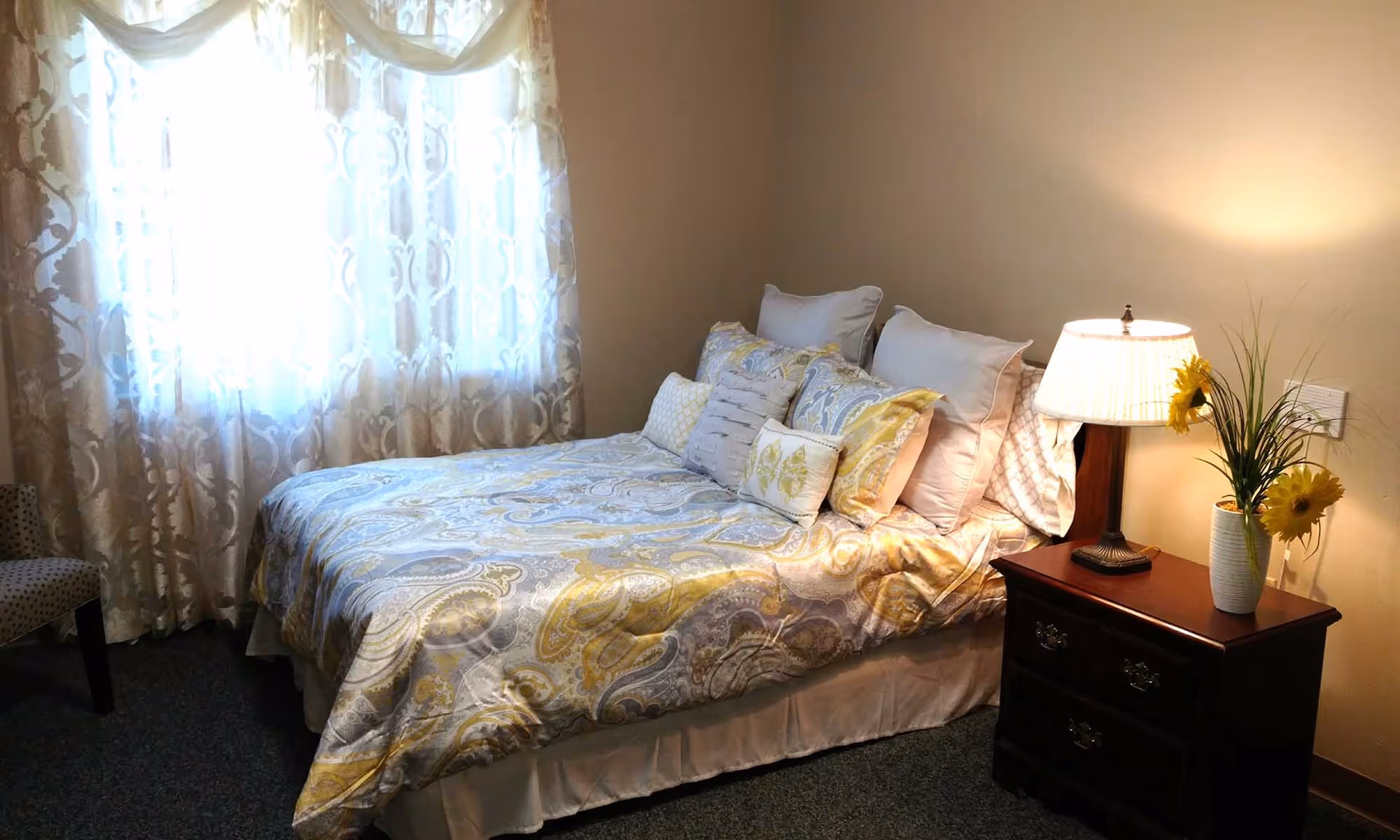 A cozy bedroom with a bed covered in a patterned yellow and gray comforter and multiple pillows. Next to the bed is a wooden nightstand with a lit table lamp and a vase with yellow flowers. Sheer patterned curtains cover a window letting in natural light. A polka-dotted chair is partially visible on the left side.