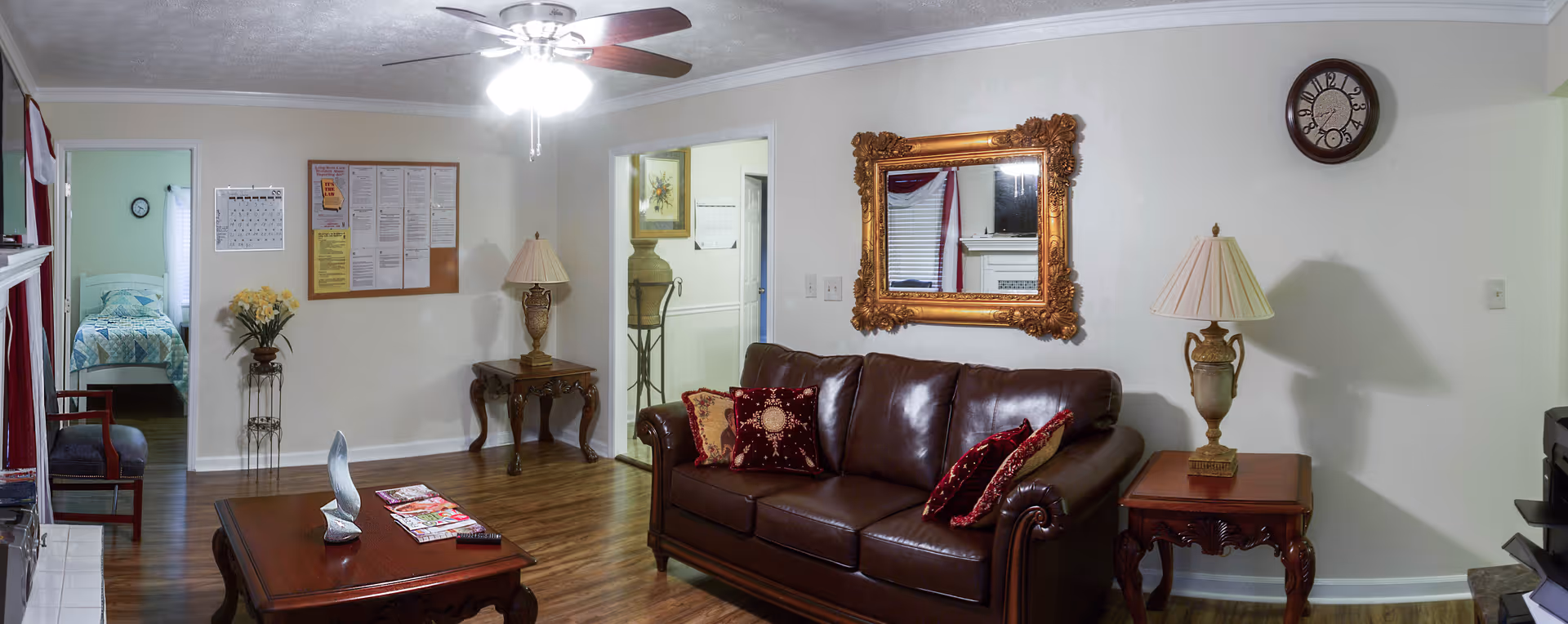 A cozy living room with a brown leather sofa adorned with red and gold decorative pillows. There is a wooden coffee table with magazines and a decorative sculpture on it. The room features wooden flooring, a ceiling fan with lights, and two side tables with ornate lamps. A large, ornate gold-framed mirror hangs on the wall above the sofa, and a round clock is mounted on the adjacent wall. In the background, there is a doorway leading to a bedroom with a bed covered in a blue and white quilt, and another doorway leading to a hallway with a decorative vase on a stand.