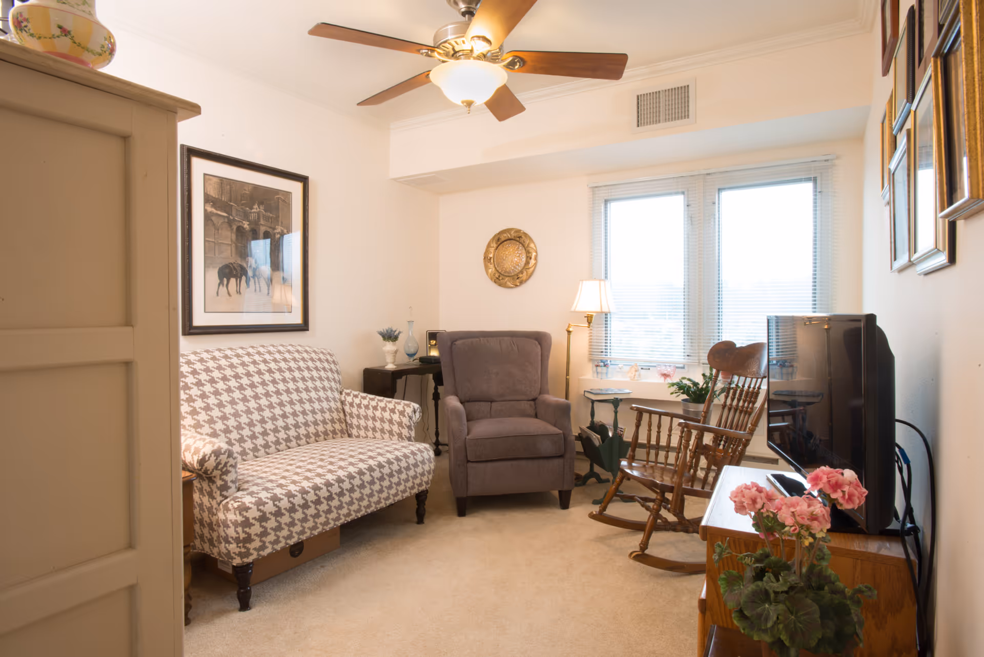 Cozy living room with a patterned loveseat, recliner, wooden rocking chair, TV, and a window with blinds.