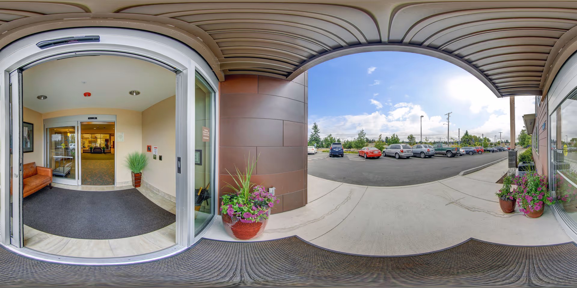 View from the entrance of Avamere Transitional Care of Puget Sound showing automatic sliding glass doors leading into a lobby area with a brown leather bench and a potted plant. Outside, there is a covered walkway with two large flower pots on either side and a parking lot with several parked cars under a partly cloudy sky.