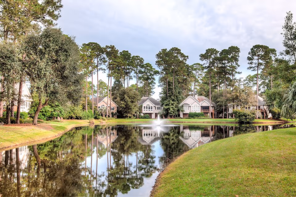 A serene outdoor scene featuring a pond with a water fountain in the center, surrounded by green grass and tall trees. Several houses are visible in the background, reflected in the calm water of the pond.