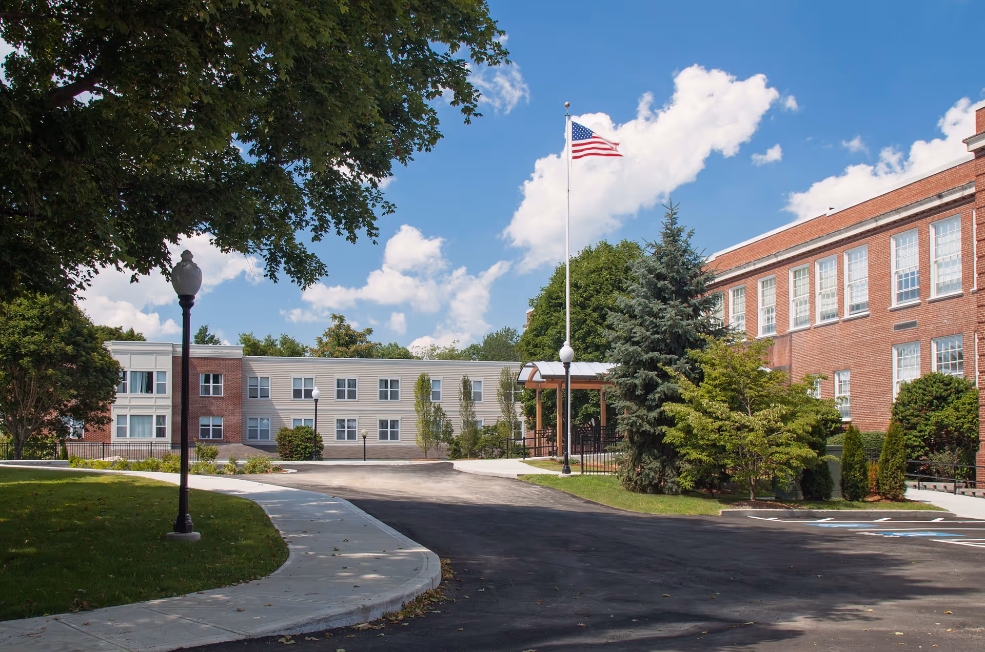 Exterior view of The Residence at Pearl Street facility showing a paved driveway, landscaped greenery, a tall flagpole with an American flag, and a two-story building with large windows under a partly cloudy sky.
