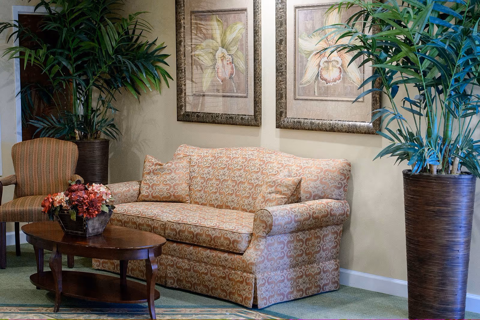 Cozy sitting area with a patterned sofa, wooden coffee table with a floral arrangement, an armchair, framed botanical prints, and large potted plants.