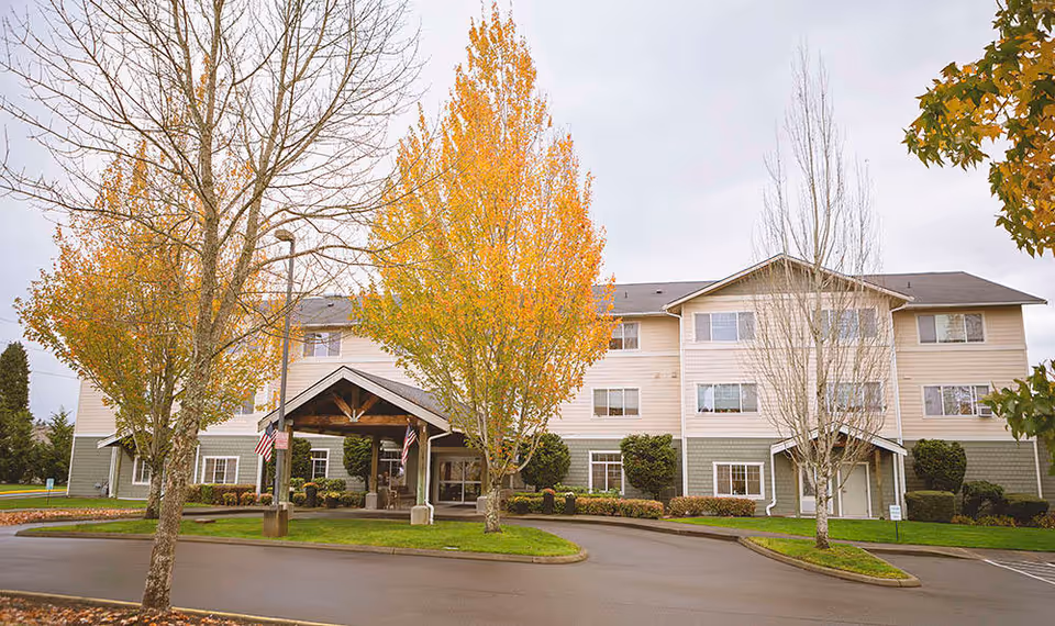 Exterior view of a three-story senior living community building with beige and green siding, surrounded by trees with autumn foliage and a driveway leading to the main entrance under a covered porch.