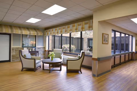 A bright and spacious common area with wooden flooring, featuring four cushioned armchairs arranged around a round coffee table with a flower vase. Large windows with patterned valances allow natural light to fill the room. The walls are painted in soft neutral tones, and there is a hallway visible to the right.