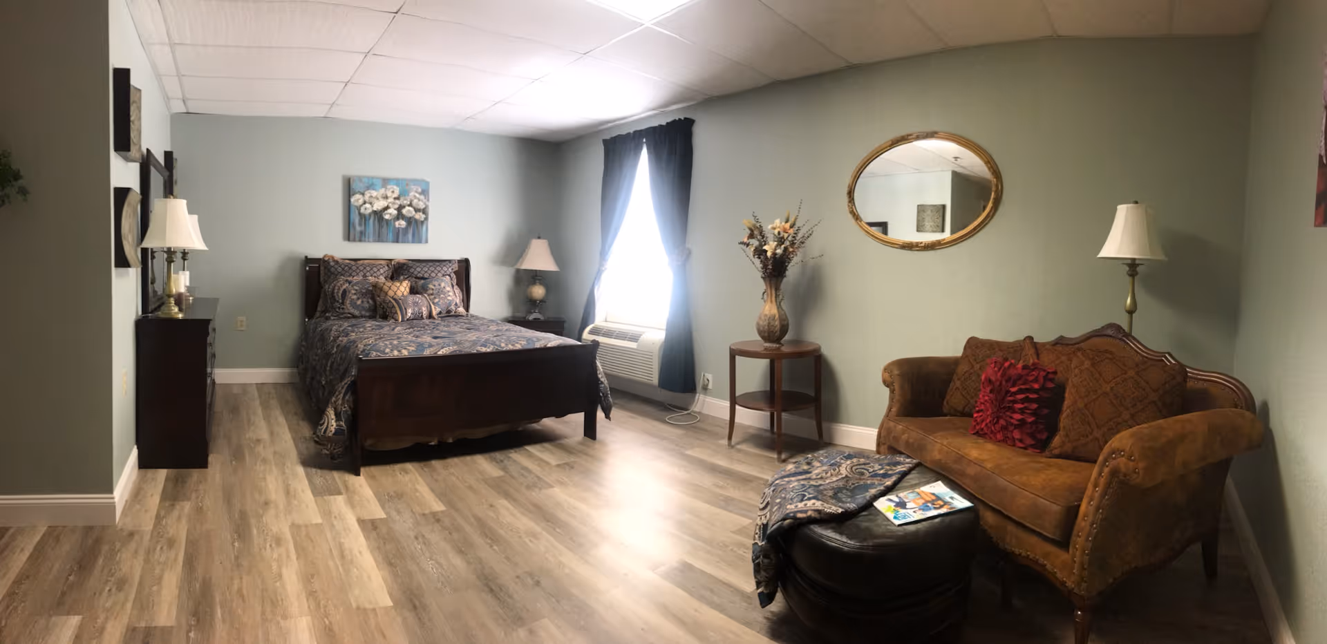 A cozy bedroom with a wooden bed featuring patterned bedding, a nightstand with a lamp, a window with dark curtains, a small round table with a vase of flowers, an oval mirror on the wall, a brown upholstered loveseat with decorative pillows, and a black ottoman with a magazine and a throw blanket on it. The room has light wood flooring and soft green walls.