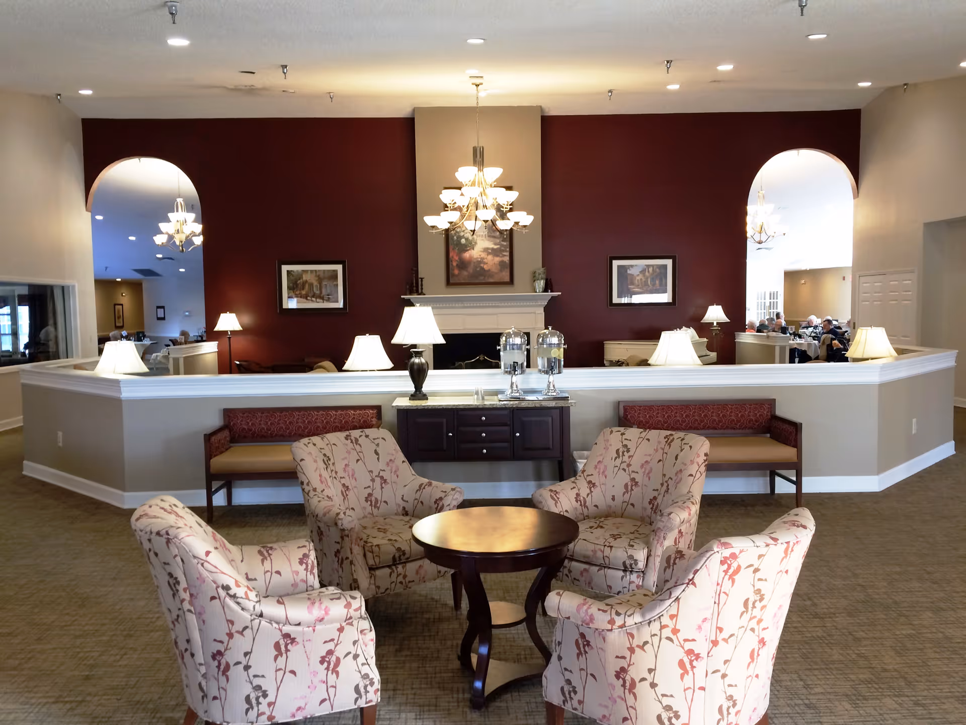Open seating area with four floral armchairs around a round wooden table facing a reception counter and chandeliers in a senior living facility.