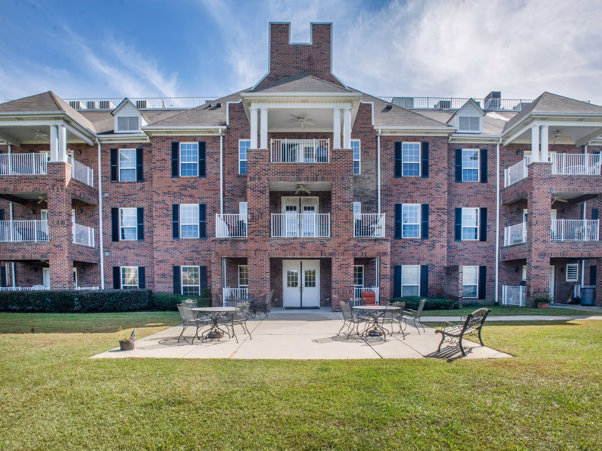 Exterior view of a three-story brick senior living facility with multiple balconies and white railings. In front of the building is a concrete patio area with metal tables, chairs, and benches on a grassy lawn under a partly cloudy sky.