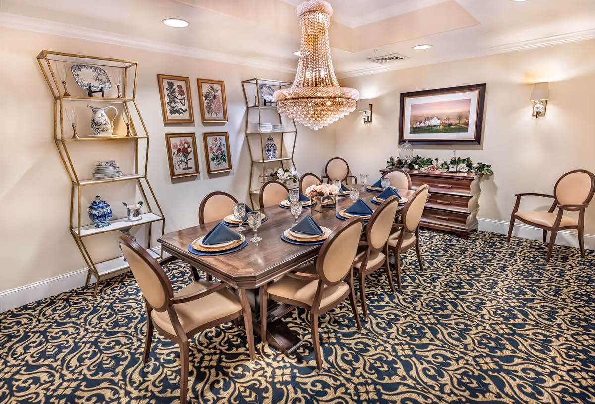 Elegant dining room with a long wooden table set for eight with plates, glasses, and navy blue napkins. The room features a large crystal chandelier, patterned carpet, two gold-framed shelves with decorative plates and vases, framed botanical prints on the wall, and a wooden sideboard with greenery and a framed landscape painting above it.