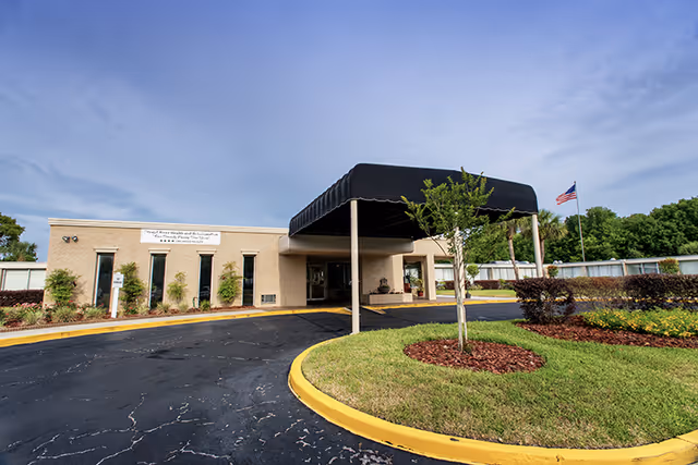 Exterior view of Crystal River Health and Rehabilitation Center showing the entrance with a black canopy, a circular driveway, landscaped greenery, and an American flag on a flagpole against a partly cloudy sky.
