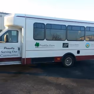 A white and maroon shuttle bus parked on a paved surface. The bus has logos and text on its side, including a green tree with the words 'Franklin Grove,' and other logos representing The Meadows of Franklin Grove Assisted Living. The text 'Proudly Serving Our Communities' is visible near the front door.