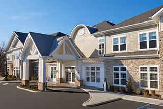 Exterior view of a two-story assisted living facility building with beige siding, stone accents, multiple windows, and a covered entrance with white columns under a blue sky.