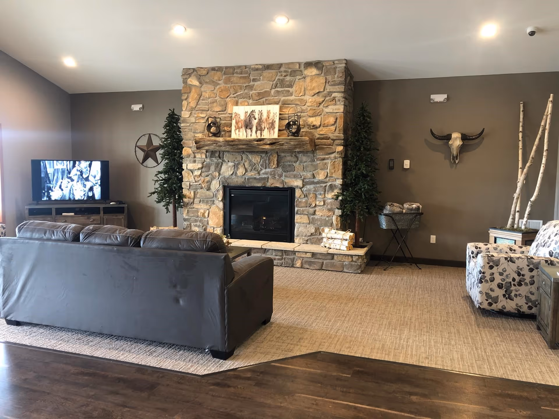 A cozy living room with a stone fireplace in the center, decorated with a painting of horses and lanterns on the mantel. There is a dark leather sofa facing the fireplace and a TV on a wooden stand to the left. The walls are painted gray and decorated with a star and a mounted animal skull. Two artificial trees flank the fireplace, and there is a patterned armchair and wooden furniture on the right side.