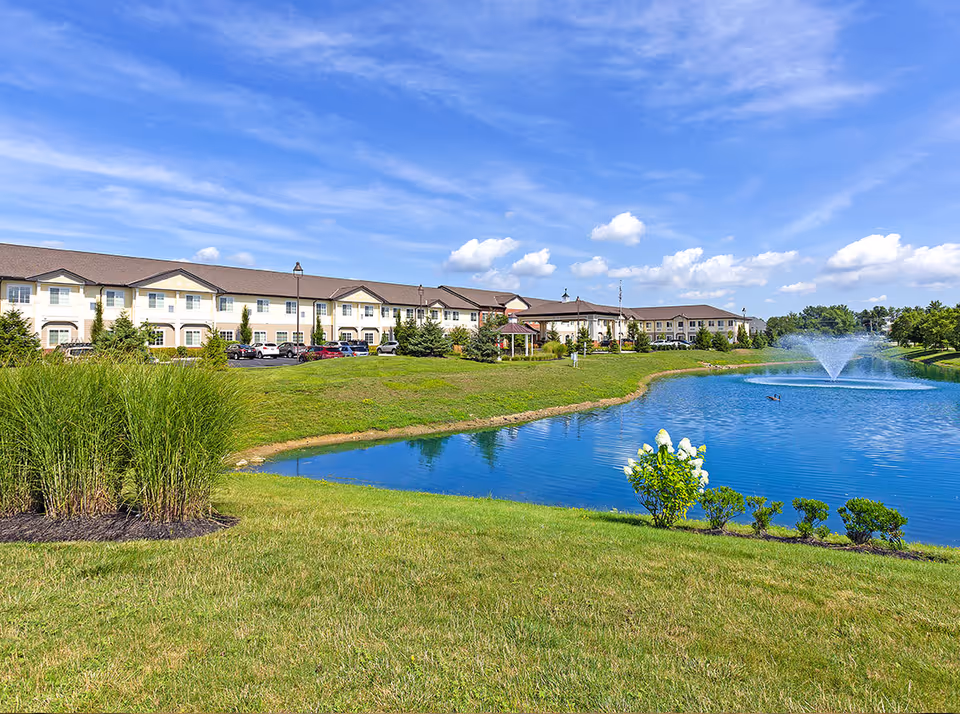 Exterior view of StoryPoint Powell senior living facility with a large pond featuring a water fountain, green grass, shrubs, and a clear blue sky with scattered clouds.