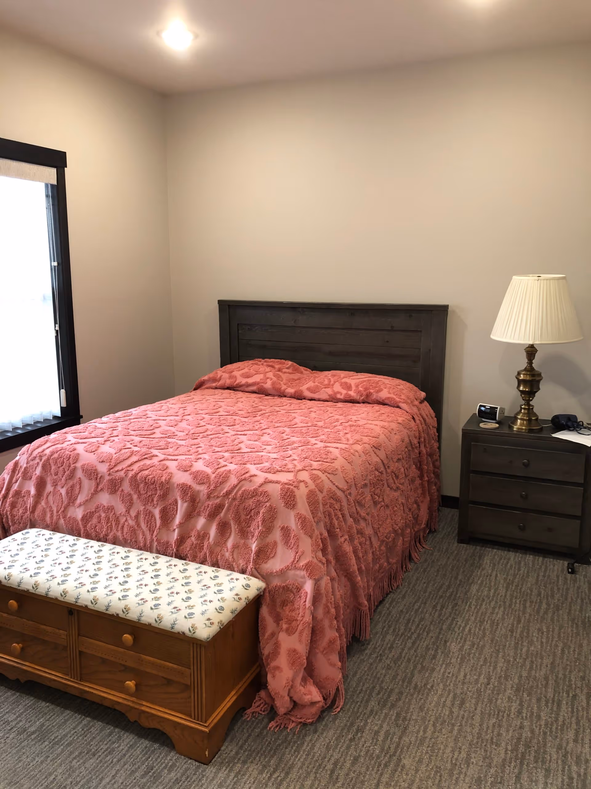 A bedroom with a bed covered in a textured pink bedspread, a wooden headboard, and a wooden bench with a floral cushioned top at the foot of the bed. There is a dark wooden nightstand with three drawers to the right of the bed, holding a brass lamp with a white lampshade, a clock, and some small items. A window with a white curtain is on the left wall, and the room has beige walls and carpeted flooring.