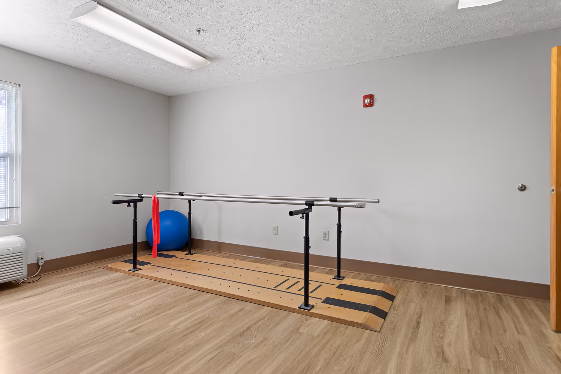 A small exercise room with wooden flooring and white walls featuring parallel bars for walking exercises, a blue exercise ball, and a red resistance band hanging on the bars. There is a window with blinds on the left and a fluorescent light on the ceiling.