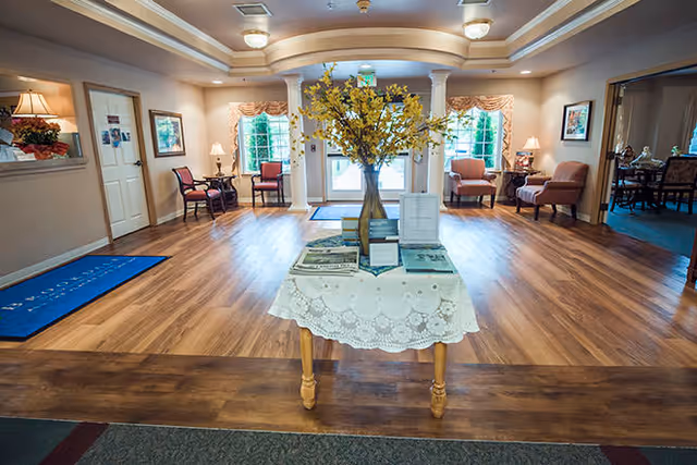 A spacious, well-lit interior common area with wooden flooring and a table in the center covered with a lace tablecloth and decorated with a vase of yellow flowers. The room features several chairs and small tables with lamps along the walls, large windows with curtains, and a double door entrance with glass panels. The ceiling has recessed lighting and decorative molding.