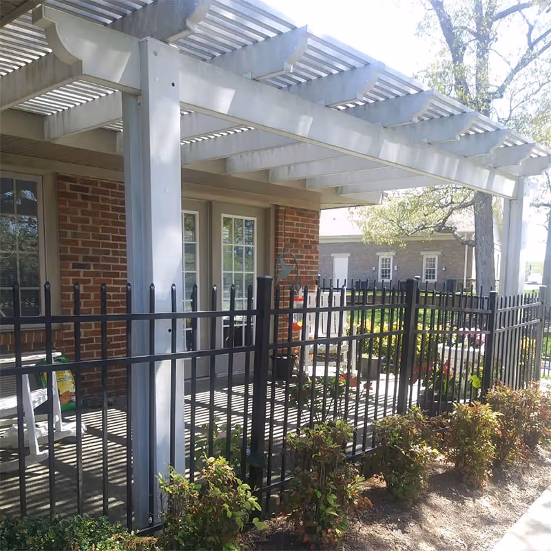 Outdoor patio area with a white pergola attached to a brick building. The patio is enclosed by a black metal fence and has some potted plants and outdoor furniture. There are bushes planted along the outside of the fence and trees in the background.
