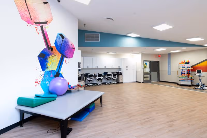 A spacious rehabilitation room with light wood flooring and white walls. On the left, there is a padded therapy table with exercise equipment including a purple exercise ball and green foam blocks. The wall behind the table features colorful geometric artwork resembling dumbbells. In the background, there are several white cabinets and a row of office chairs along a counter. On the right side, there is exercise equipment including a stationary bike and a rack with various colorful weights. The room is well-lit with ceiling lights and has a modern, clean appearance.