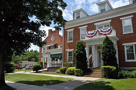Brick colonial-style senior living building with patriotic bunting and an American flag at the front entrance, surrounded by trees and a sidewalk.