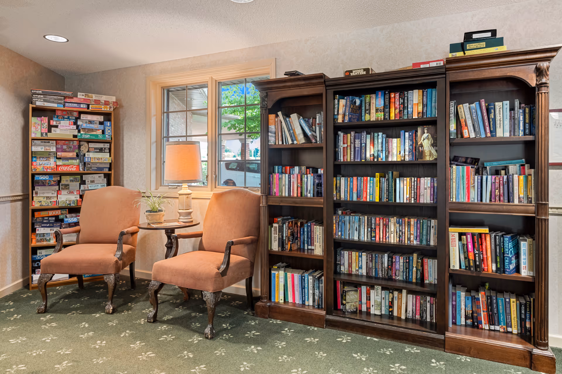 Cozy reading area with two upholstered chairs, a side table and lamp, and large bookshelves filled with books and board games nearby.